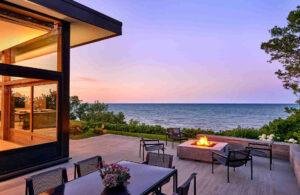 Modern patio with a fire pit and outdoor dining set overlooks a calm body of water at sunset, bordered by greenery and trees.