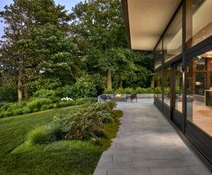A modern home with large glass walls opens onto a sleek stone patio surrounded by lush landscaping. The patio features a seating area with black chairs and a linear fire pit. The surrounding garden is filled with ornamental grasses, shrubs, and flowering plants, with tall mature trees providing a green backdrop.