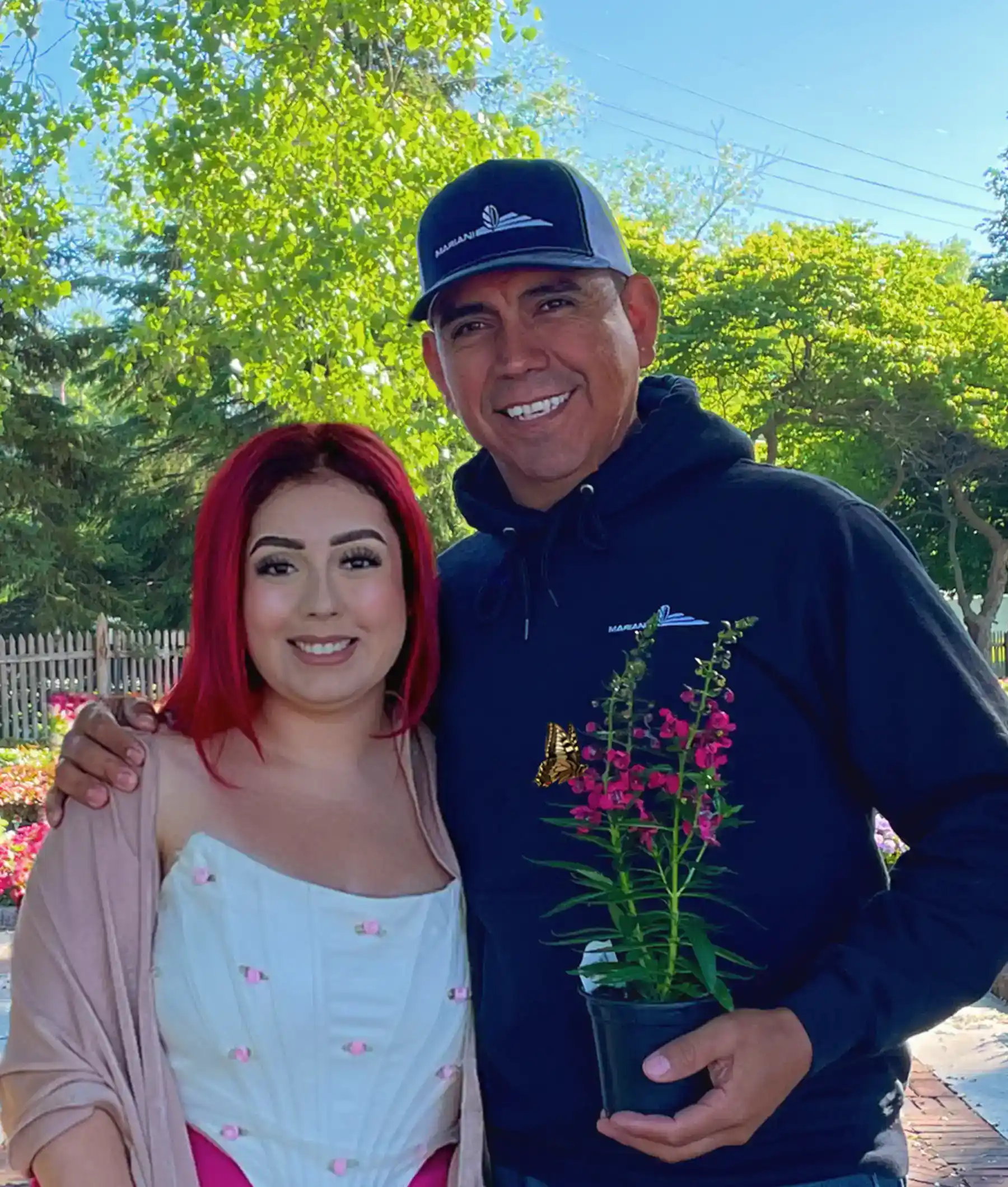 A smiling man and woman stand close together outdoors. The man holds a small potted plant. The woman has red hair and wears a white dress. Trees and a wooden fence are visible in the background.