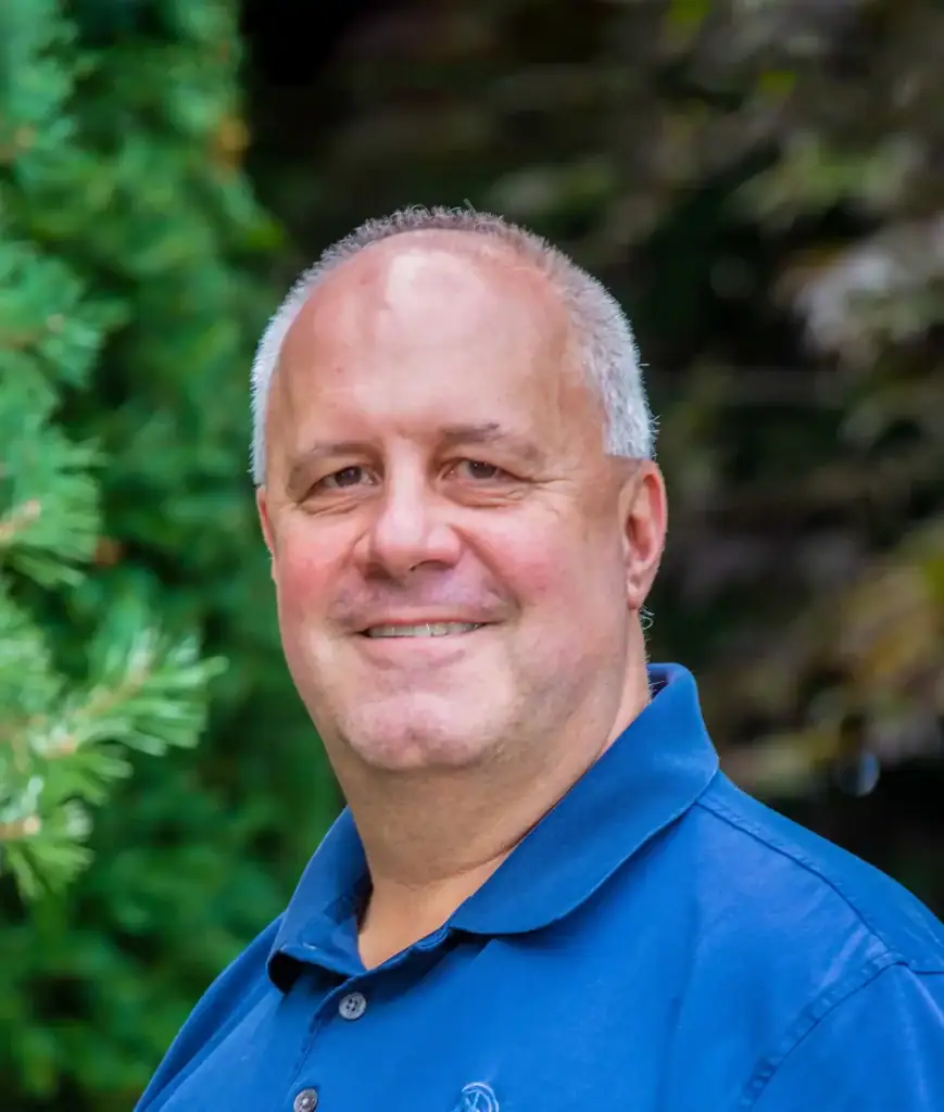 A man with short grey hair and a blue polo shirt smiles while standing outdoors in front of green foliage.