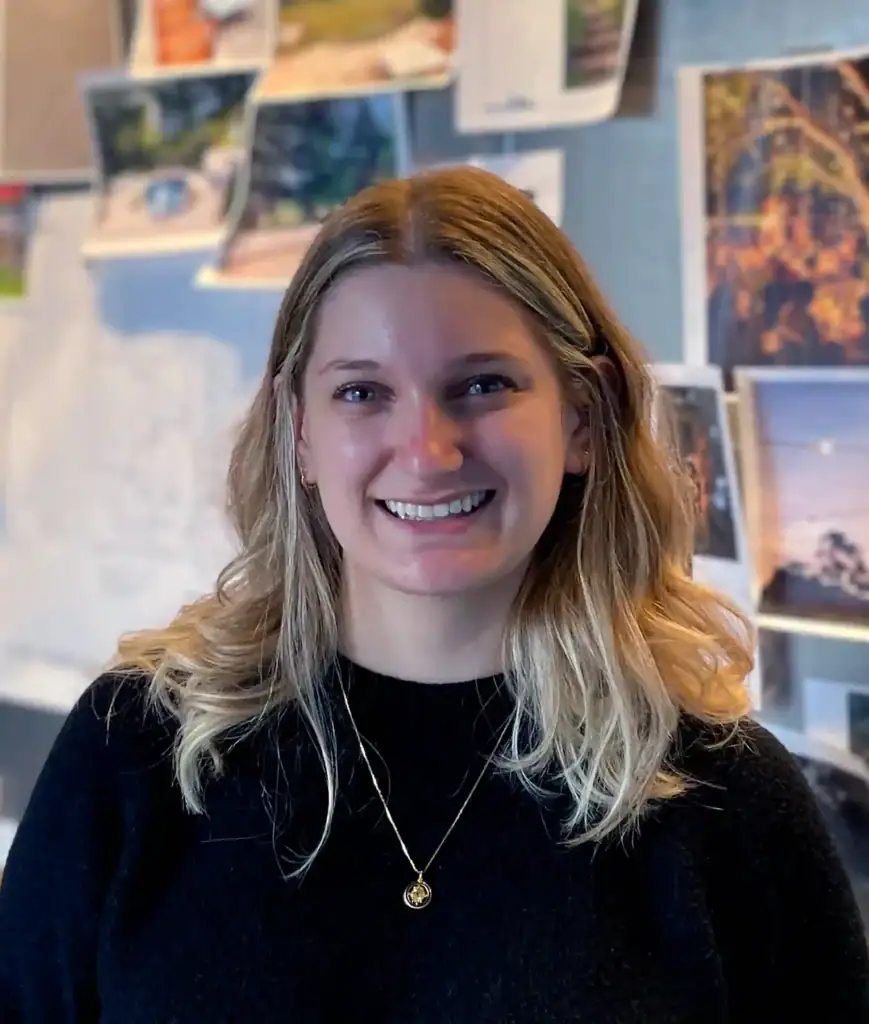 A woman with long blonde hair smiles while standing in front of a wall covered with various photos and papers. She is wearing a black top and a gold pendant necklace.