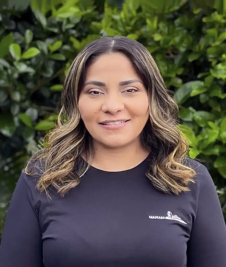 A woman with wavy hair, wearing a black shirt, stands in front of a background of green foliage.