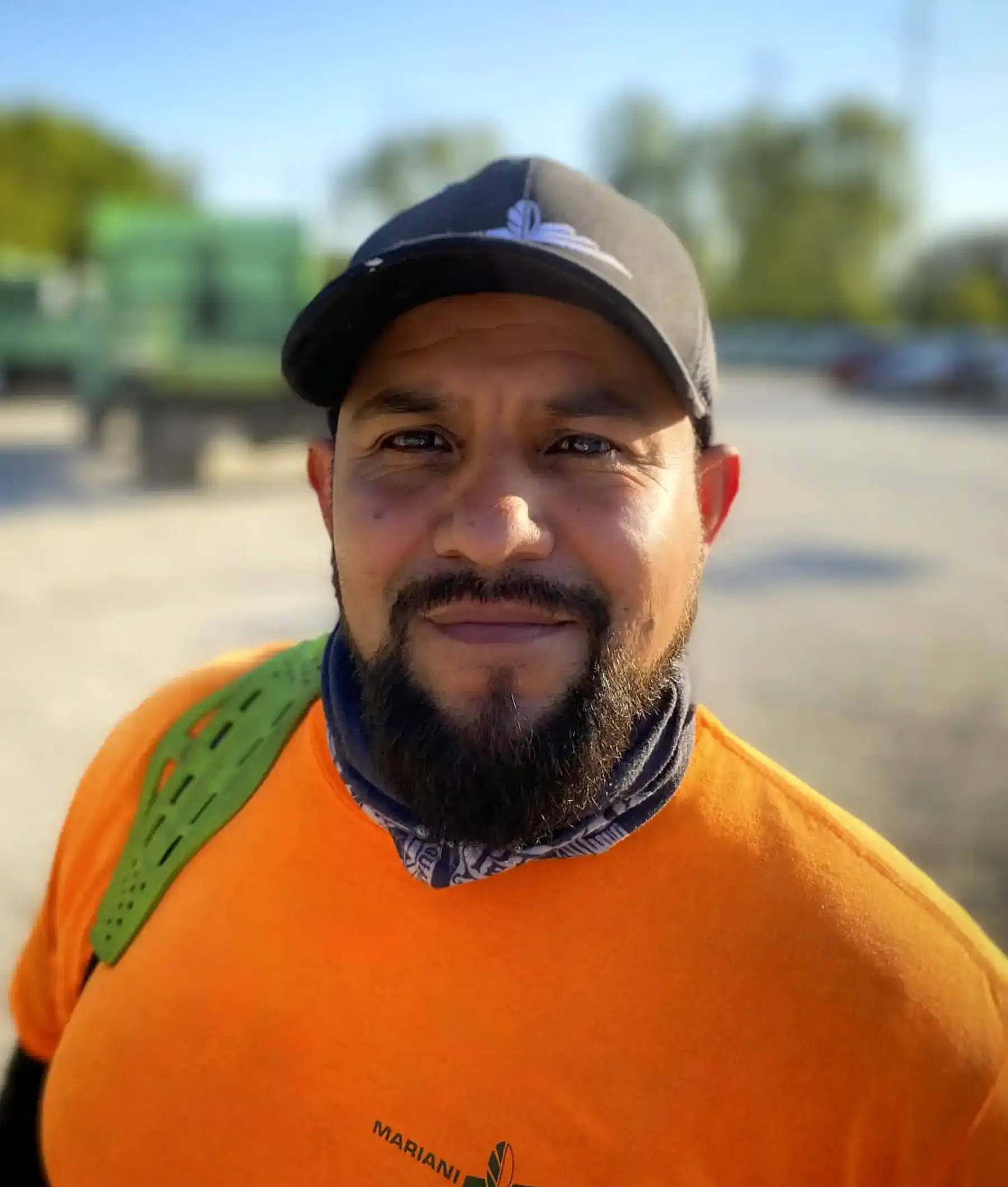 A man with a beard, wearing an orange shirt and black cap, smiles at the camera outdoors. The background is blurred, showing green machinery and trees.