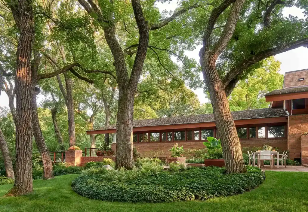 A modern, single-story brick house is surrounded by tall trees and lush green grass, featuring large windows and a small outdoor seating area.