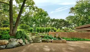 A landscaped garden featuring stone steps, rock formations, a small fountain, and lush greenery alongside a brick structure under a partly cloudy sky.