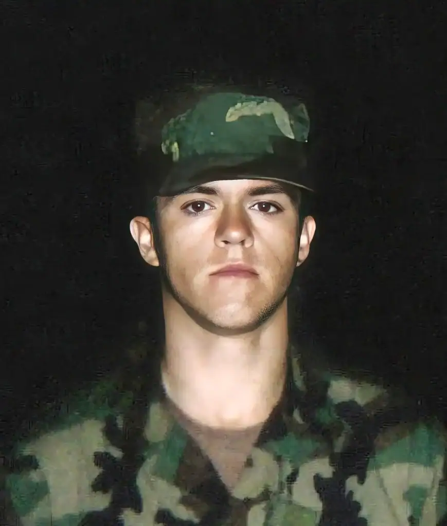 A person wearing a camouflage military uniform and hat, posing for a portrait against a dark background.