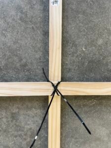 A wooden frame with two black zip ties securing the intersecting beams on a gray stone floor.