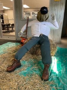 A scarecrow dressed in protective gear, sitting on hay inside a room, with a hedge trimmer by its side.