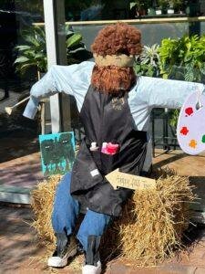Scarecrow dressed as an artist, seated on hay, wearing an apron. It holds a palette and brushes, with a canvas painting nearby. A small sign reads "happy little trees.