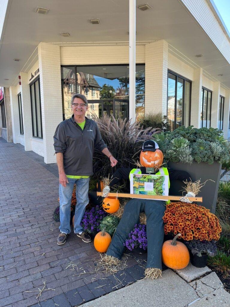 A man stands next to a pumpkin scarecrow display surrounded by plants and pumpkins outside a building.