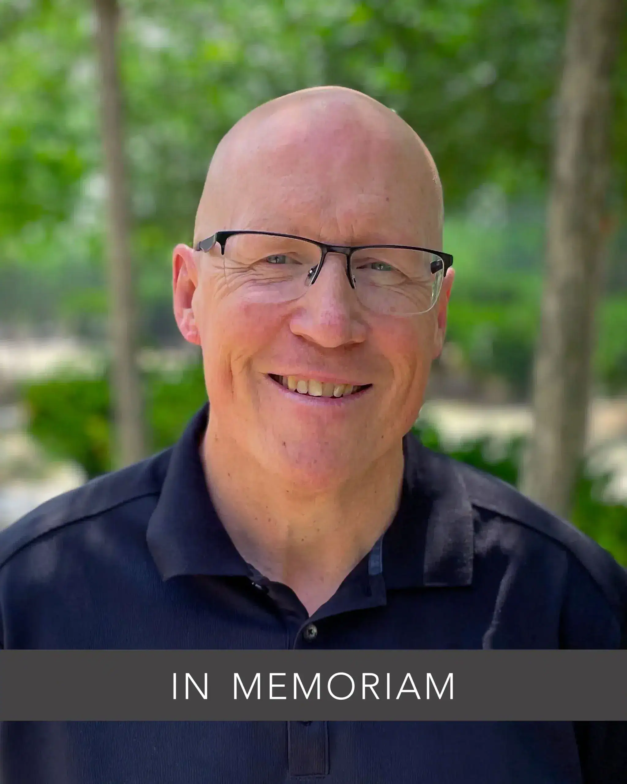 Bald man with glasses wears a black polo shirt, smiling outdoors in a leafy setting. Text reads "In Memoriam.