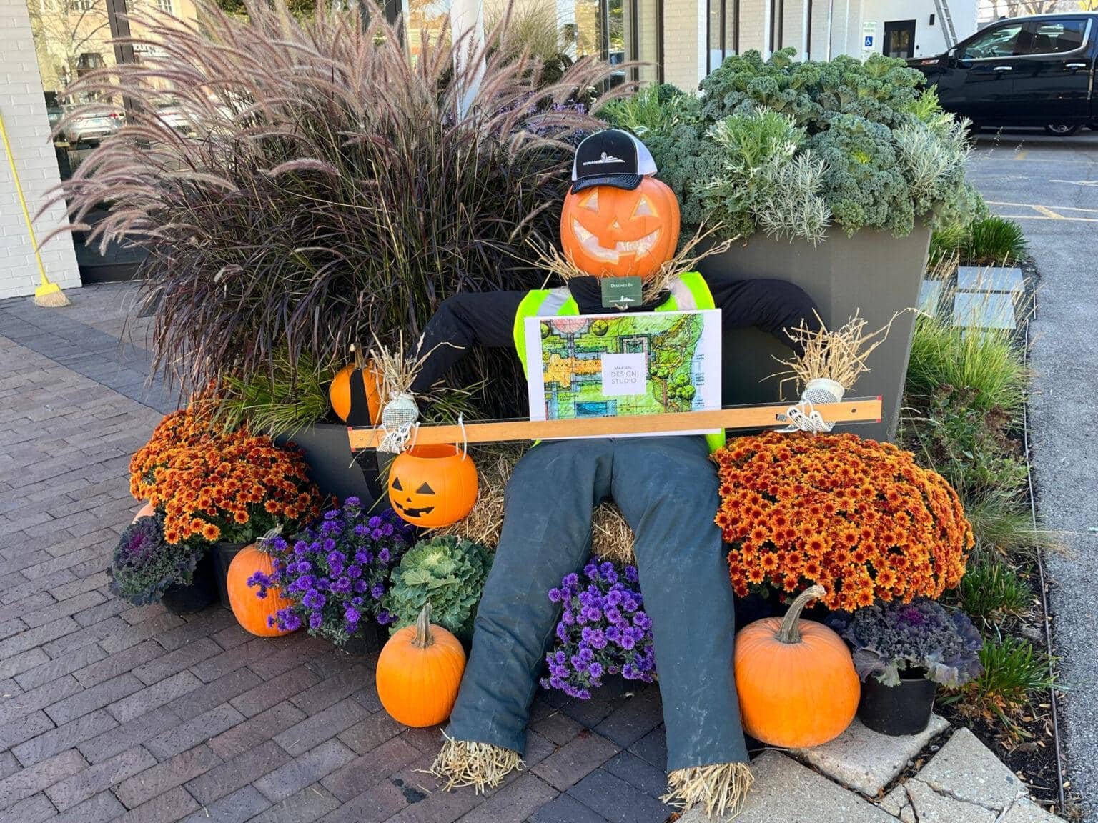 A scarecrow with a pumpkin head sits among vibrant orange and purple flowers, surrounded by pumpkins, on a brick pavement near a building.