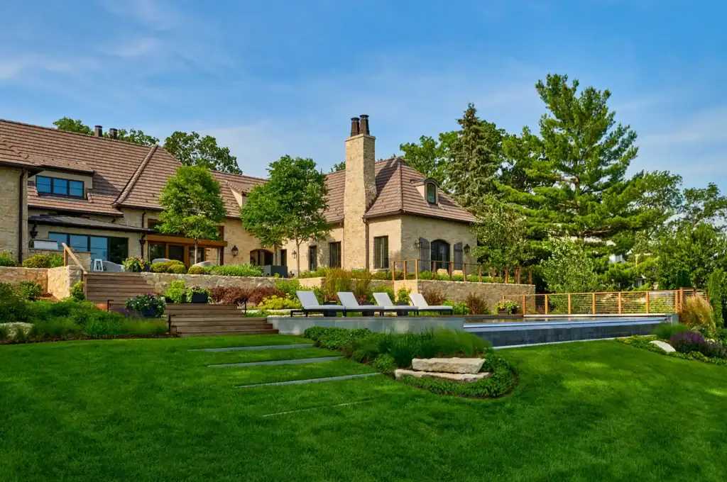 Spacious stone house with landscaped garden, pool, and several lounge chairs on a sunny day. Trees and blue sky in the background.