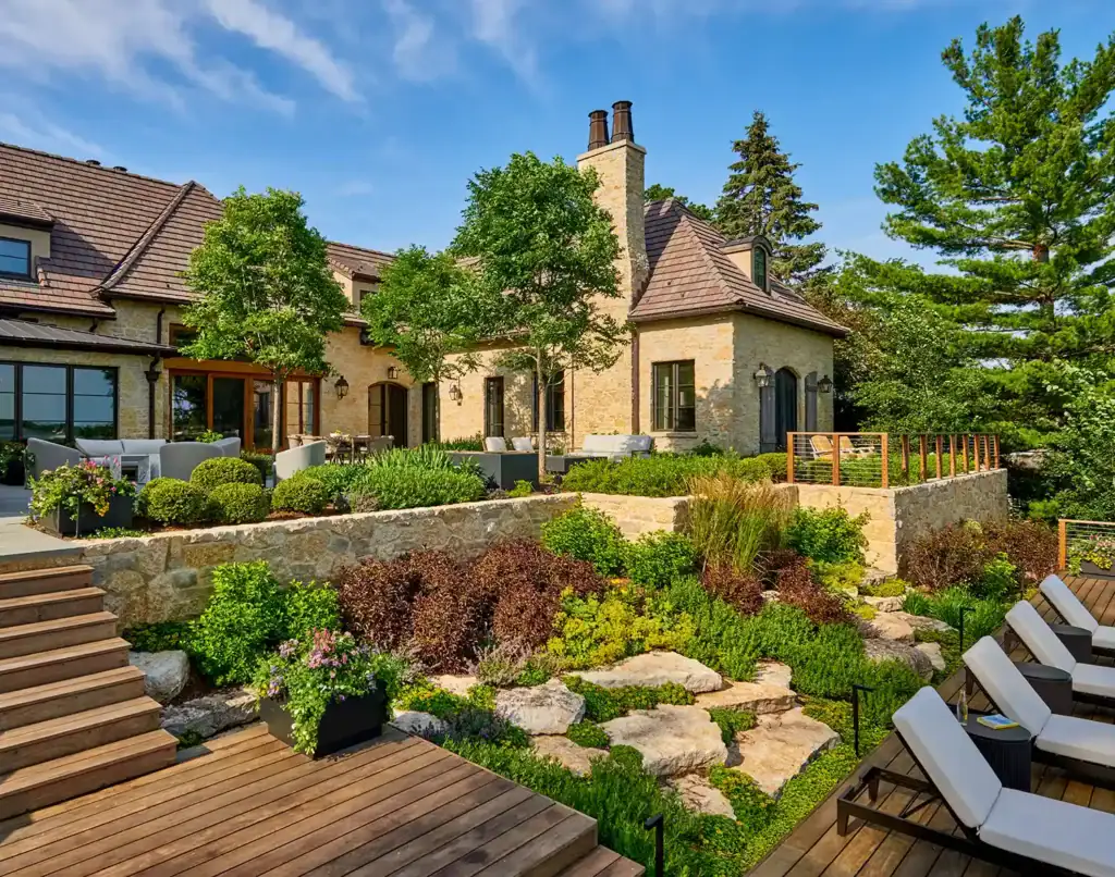 Stone house with sloped roof, surrounded by trees and landscaped garden. Wooden deck with lounge chairs in foreground under a clear blue sky.