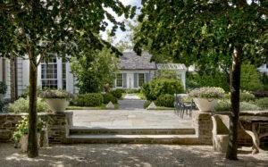 A garden with stone steps leading to a patio, surrounded by lush greenery and large potted plants. A table and chairs are set on the right side under trees.