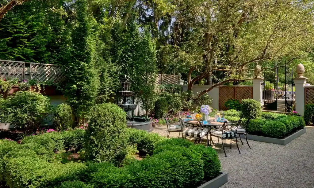 A garden with a gravel path, manicured bushes, patio furniture, a dining table with decor, and a tiered fountain surrounded by trees and lush greenery.