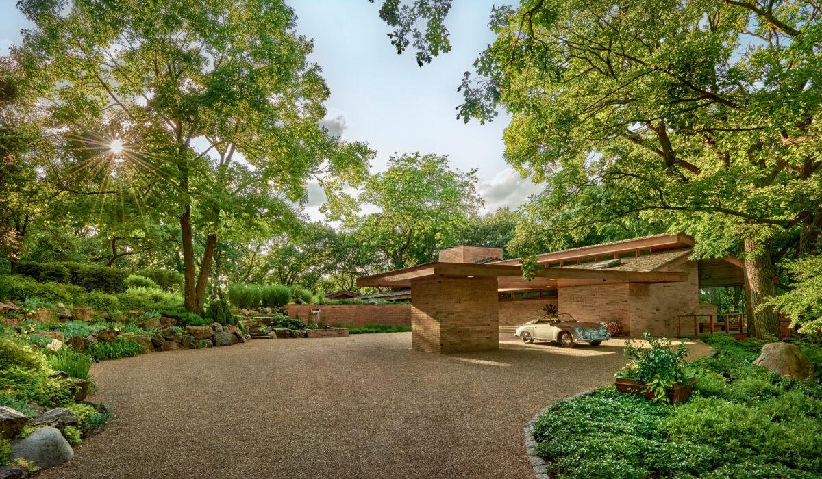 A modern house with flat roofs and large windows is surrounded by lush trees and a garden. A car is parked in the gravel driveway. Sunlight filters through the foliage.