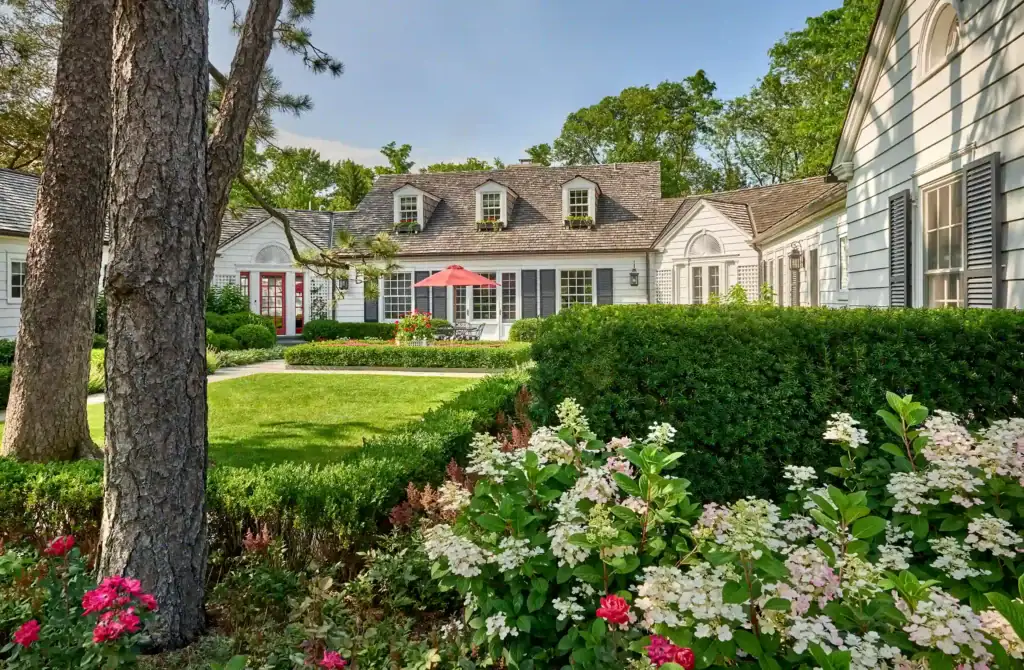 Charming white house with gray shutters, surrounded by lush greenery and flowers. Manicured lawn, red patio umbrella, and clear blue sky.