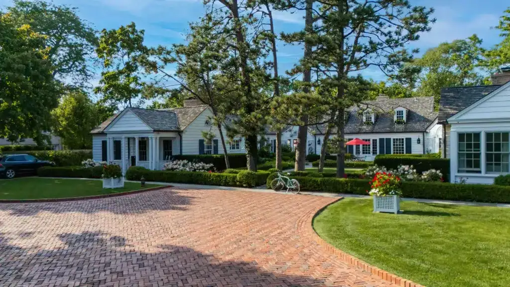 A brick driveway leads to a large white house surrounded by trees and landscaped gardens. A bicycle is parked in front, and a red umbrella is visible in the backyard.