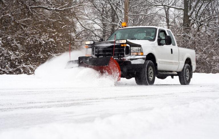 A white truck with a snowplow attachment efficiently handles snow and ice removal for hospitals, ensuring clear pathways. In the background, snow-covered trees create a serene winter landscape.