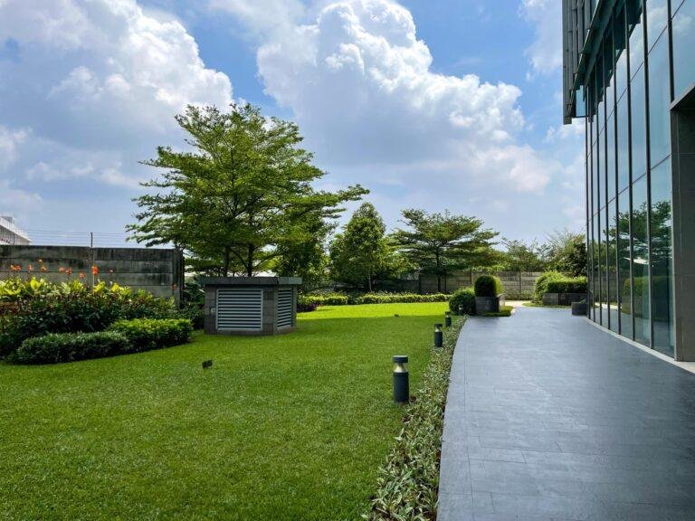 Modern building exterior with glass facade, adjacent to a meticulously landscaped garden maintained for hospital standards, featuring lush green grass, shrubs, and trees under a partly cloudy sky.