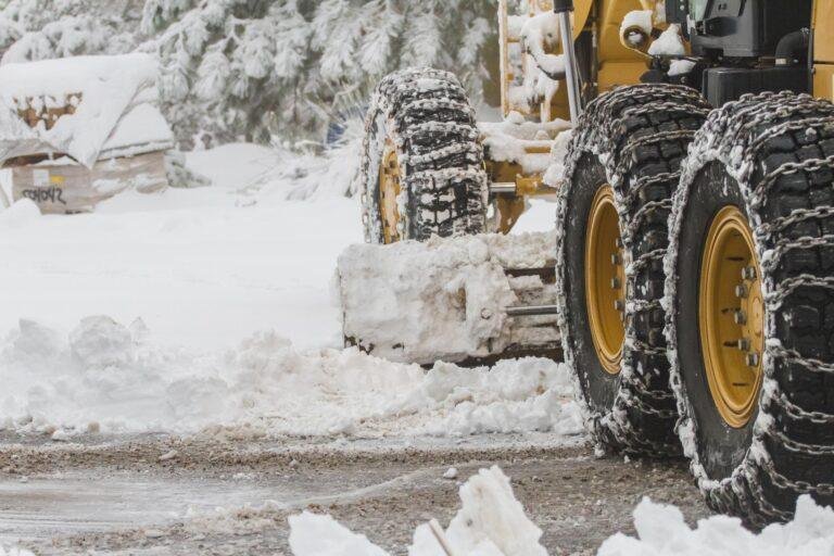 A snowplow with chained tires expertly handles snow and ice removal for large venues, clearing a road in a snowy, forested area.