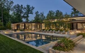 Modern house exterior at dusk with large windows, a rectangular swimming pool, poolside lounge chairs, and landscaped greenery.