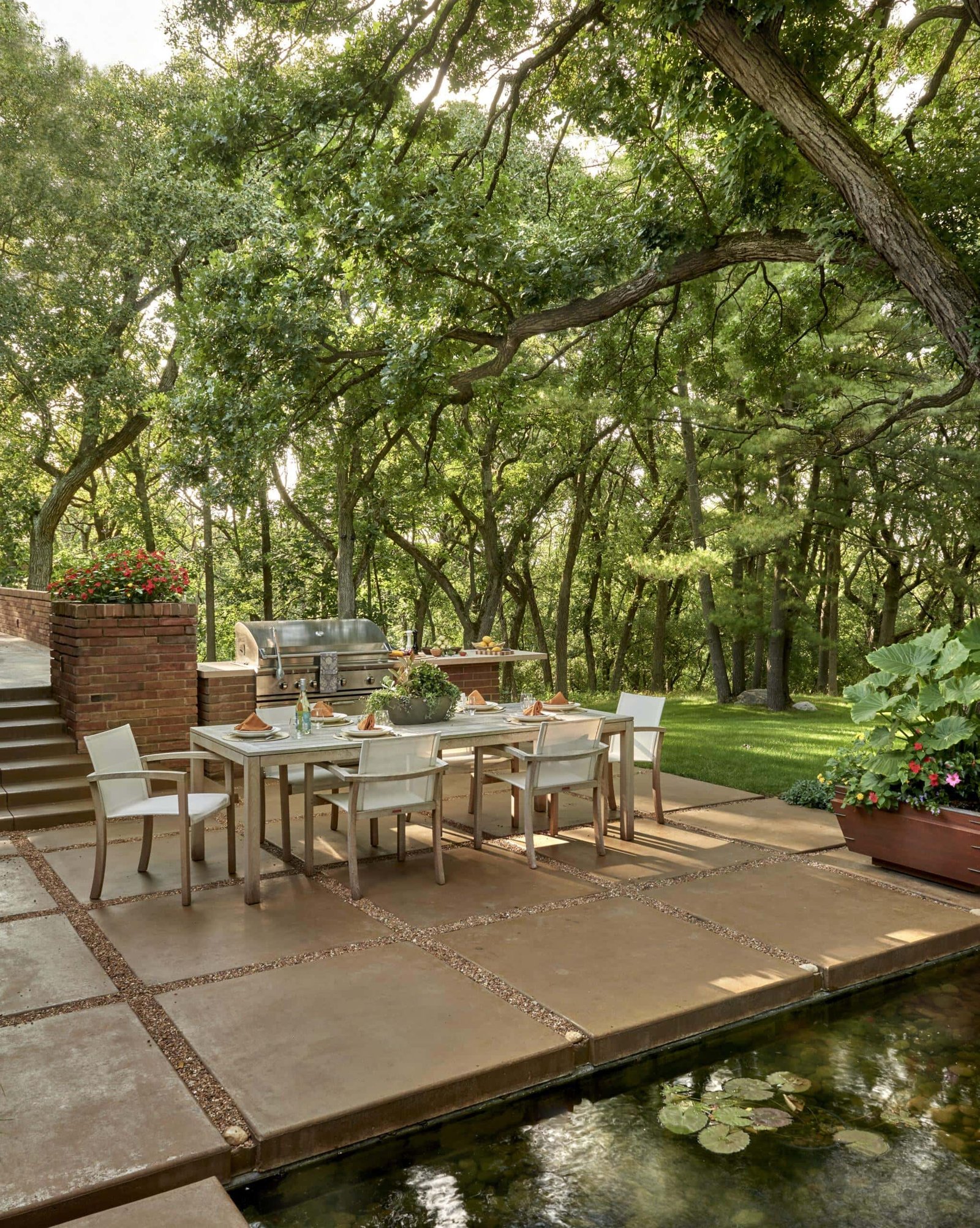 Outdoor dining area with a modern table and chairs on a patio, surrounded by lush trees and expert landscaping in Barrington IL, next to a small pond with lily pads and a built-in grill.