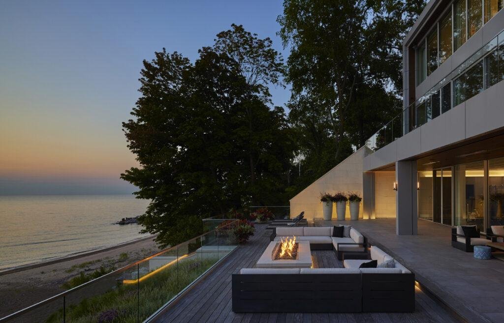 Modern outdoor patio with a fire pit, seating area, and glass railing overlooks a beach and water at sunset, next to a contemporary house with large windows.