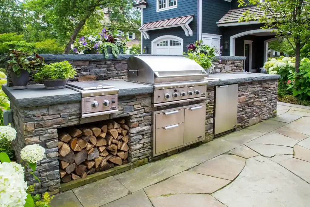 Outdoor stone kitchen with built-in stainless steel grill, stove, storage, and a wood compartment, surrounded by plants and paving stones.
