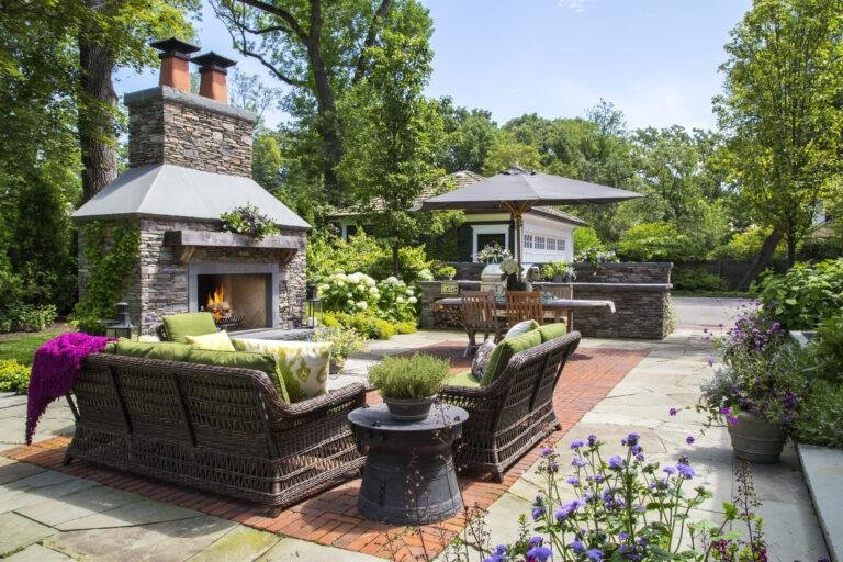 Outdoor patio area with wicker furniture, cushions, a stone fireplace, dining table with umbrella, and lush garden plants—showcasing expert landscaping in Hinsdale, IL amid trees on a sunny day.