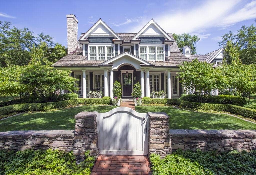 Two-story house with gray siding, large windows, and a front porch, featuring manicured landscaping in Hinsdale, IL, all enclosed by a stone fence with a white gate.