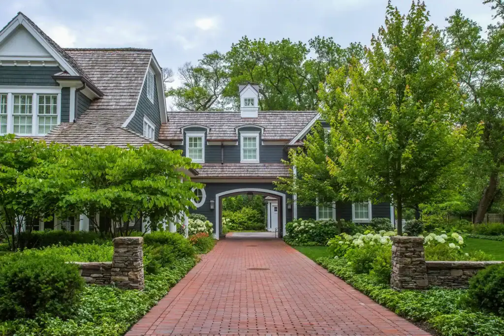 A two-story gray house with white trim, surrounded by green trees and shrubs, features a red brick driveway and stone pillars at the entrance.