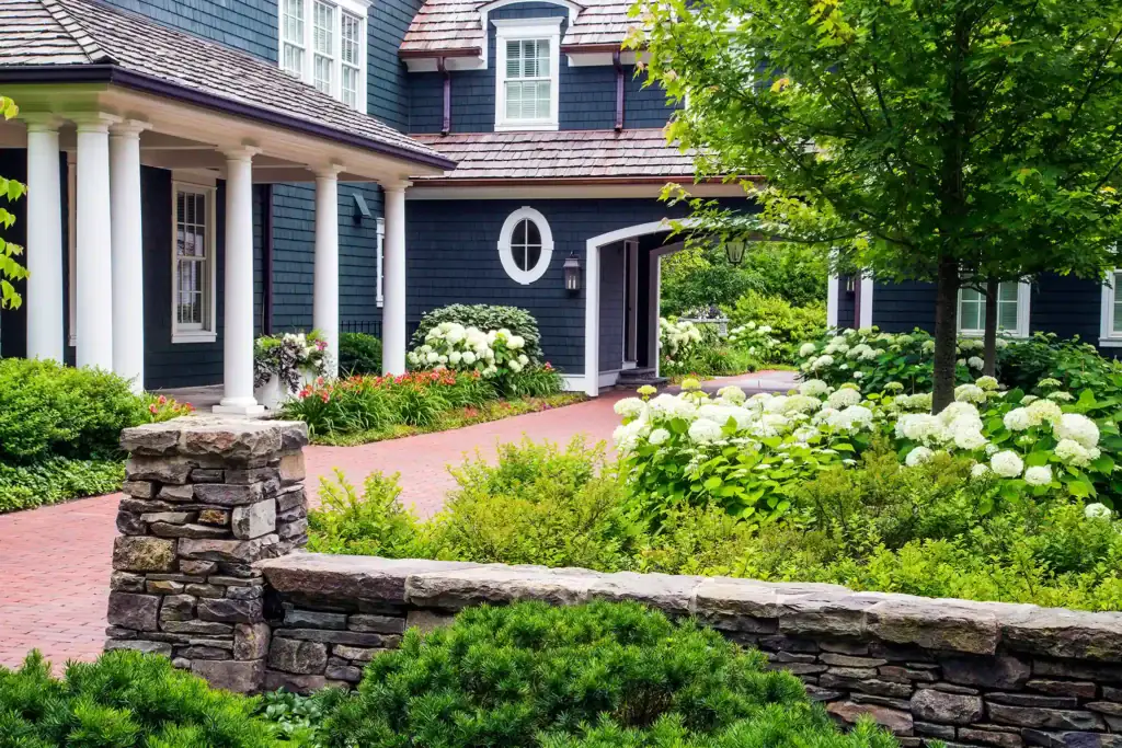 A well-maintained house with dark blue siding, white trim, and a brick pathway bordered by lush landscaping, hydrangeas, and a stone wall in the foreground.