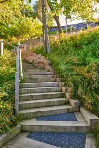 Wooden steps with gravel sections ascend a grassy hillside, bordered by railings and surrounded by green foliage and trees on a sunny day.