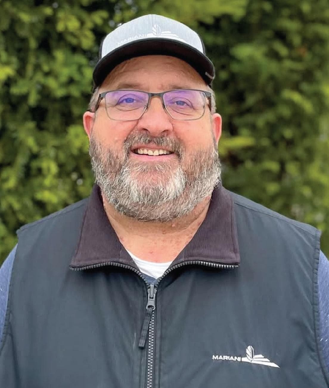 A man with a gray beard and glasses, wearing a cap and a black vest, stands outdoors in front of green foliage.