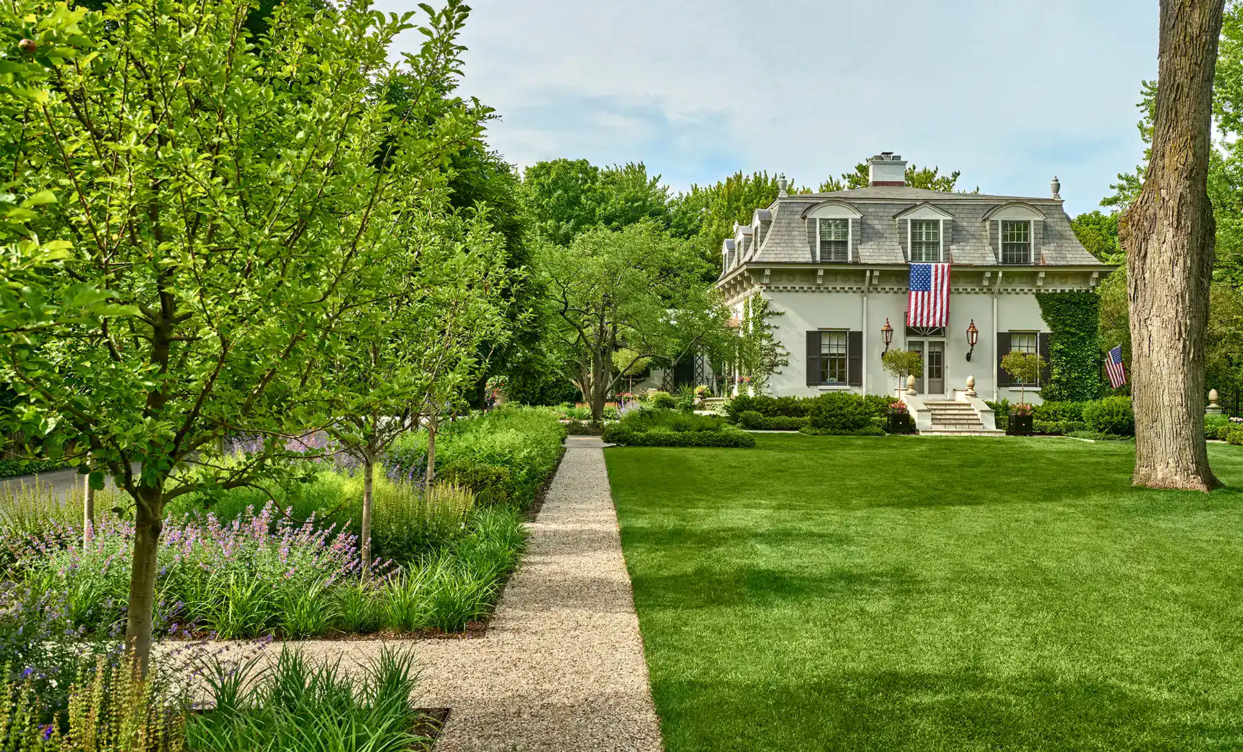 A well-manicured lawn with a gravel path leads to a white two-story house displaying American flags and surrounded by lush trees and gardens.