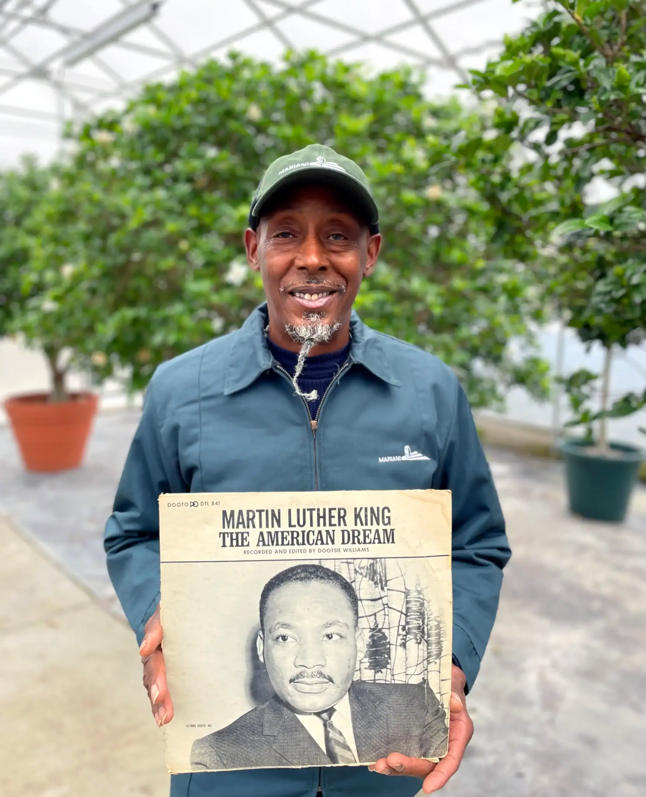 A man in a blue jacket and green cap stands in a greenhouse holding an album titled "Martin Luther King: The American Dream" with a photo of Martin Luther King Jr. on the cover.