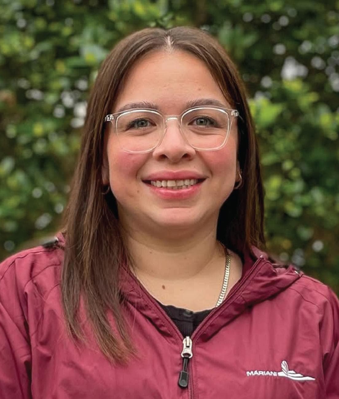 A woman with straight brown hair, glasses, and a maroon jacket smiles in front of green foliage.