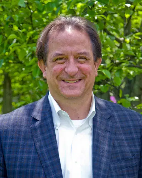 A man with brown hair wearing a white shirt and a blue plaid blazer stands smiling outdoors in front of green foliage.