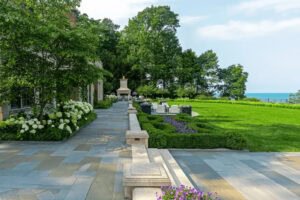 A landscaped backyard with stone patio, manicured lawn, garden beds, outdoor seating area, fireplace, and a view of trees and water in the background.