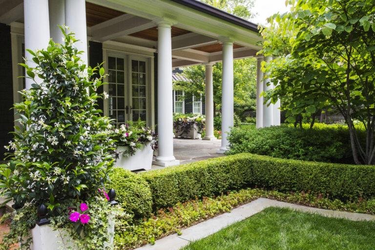Front porch of a house with white columns, potted plants, manicured hedges, flowering shrubs, and a neatly trimmed green lawn.