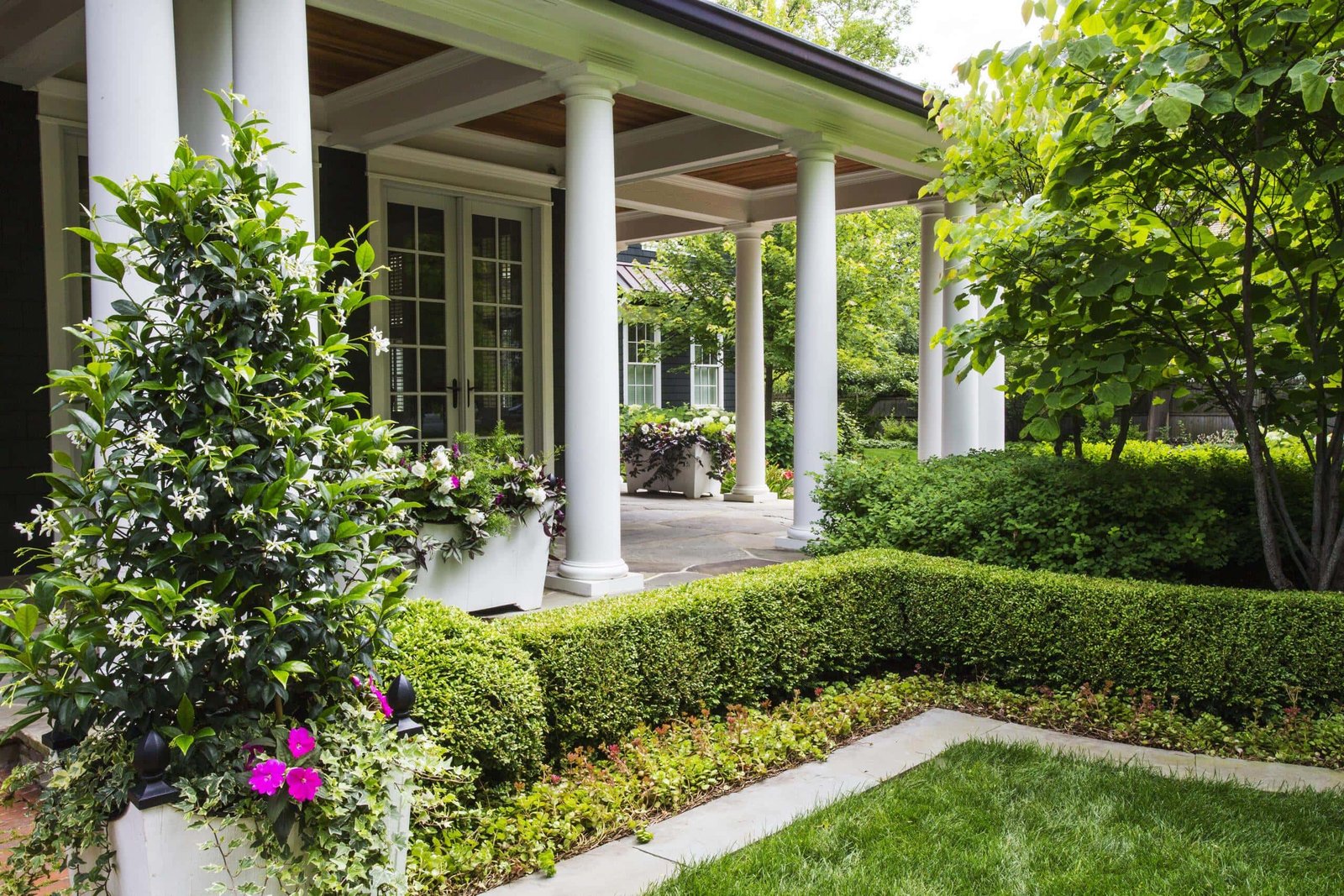 Front porch of a house with white columns, potted plants, manicured hedges, flowering shrubs, and a neatly trimmed green lawn.