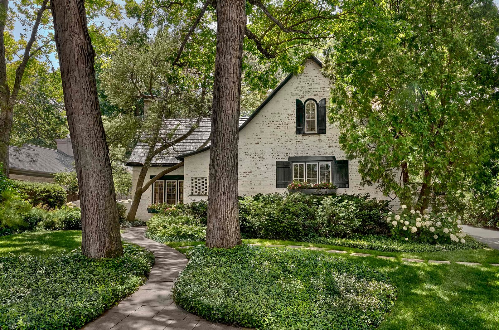 A white brick house with black shutters, arched windows, and a steep roof surrounded by mature trees, lush greenery, and a curved stone pathway.