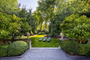 A landscaped garden with green lawn, three lounge chairs, stone planters, and surrounding trees and shrubs under clear daylight.